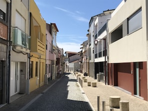 A narrow cobblestone street lined with multi-story buildings, some with balconies and storefronts. - Povoa do Mar (Povoa De Varzim)