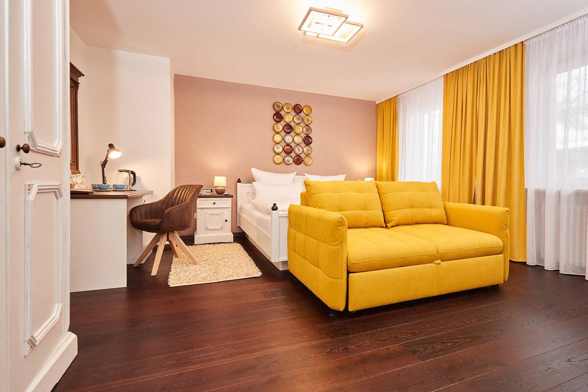 A modern bedroom with a yellow sofa, a wooden chair, a bedside table with a lamp, and a wall-mounted plate decoration.