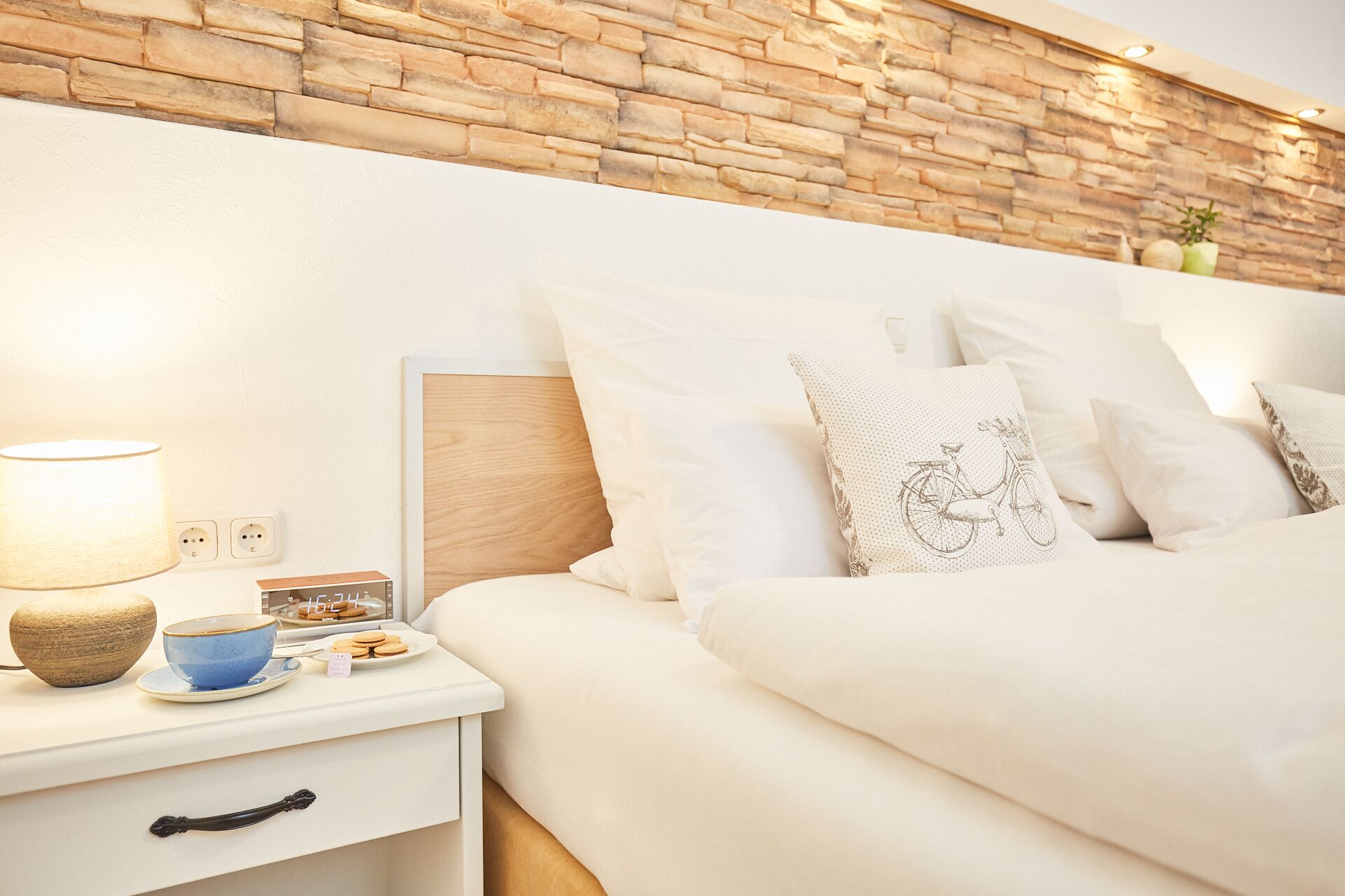 A neatly made bed with white bedding, a wooden headboard, and a nightstand with a lamp, a cup, and cookies.