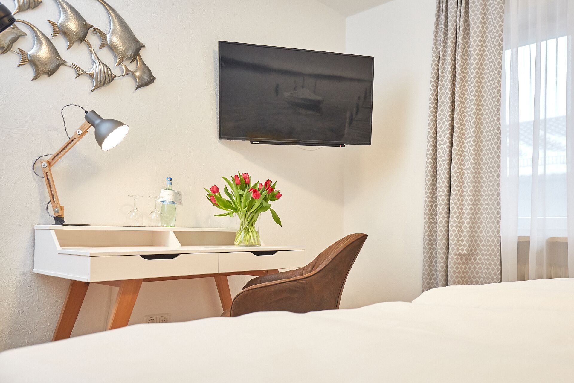 Habitación de hotel con cama, escritorio con lámpara, televisión, florero con flores y ventana con cortinas.
