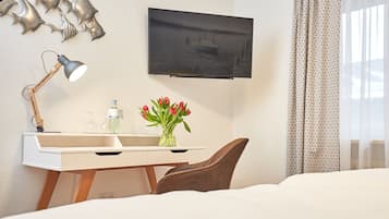 Habitación de hotel con cama, escritorio con lámpara, televisión, florero con flores y ventana con cortinas.