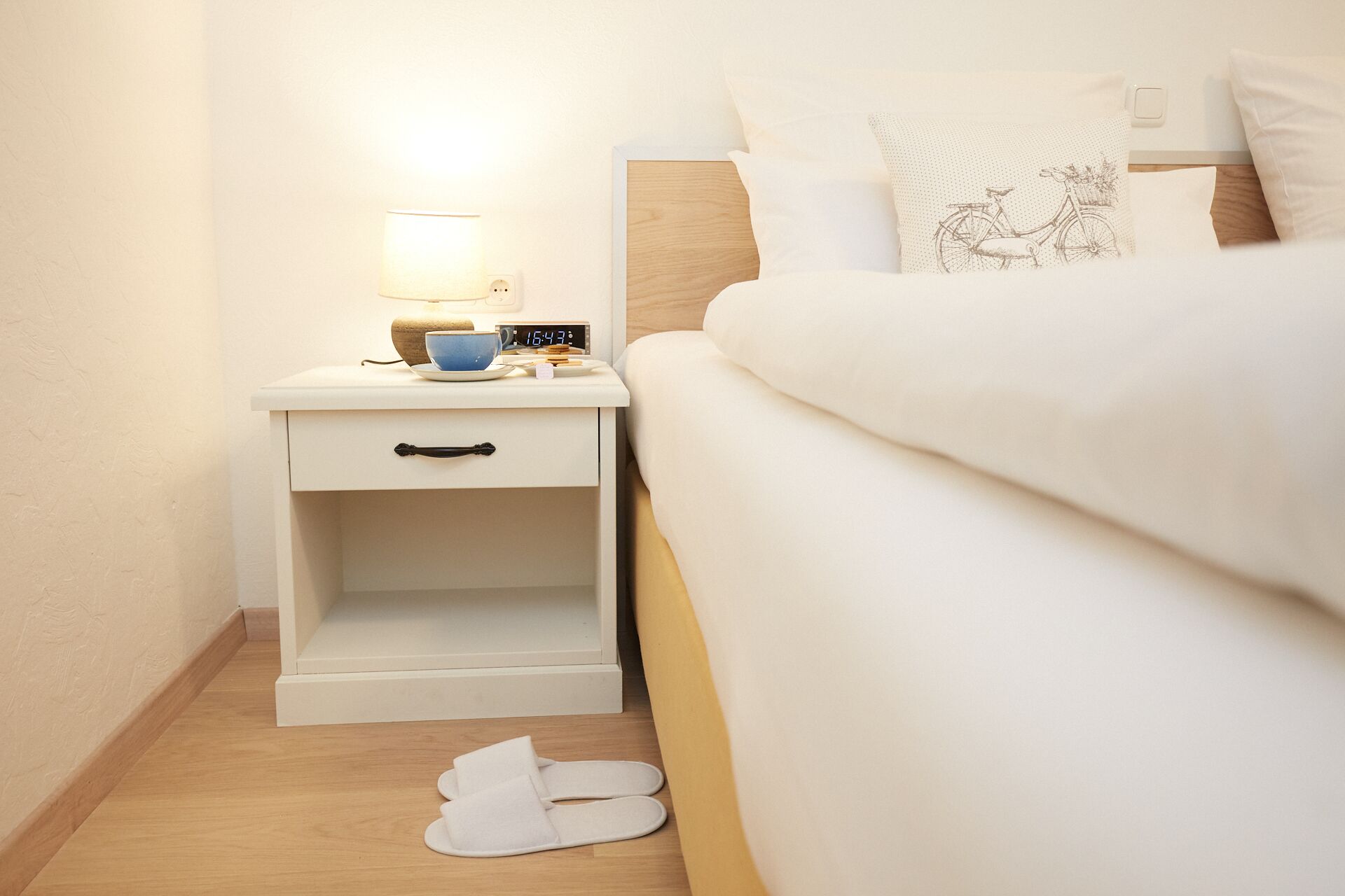 A neatly made bed with white bedding, a bedside table with a lamp, a clock, and a blue cup.