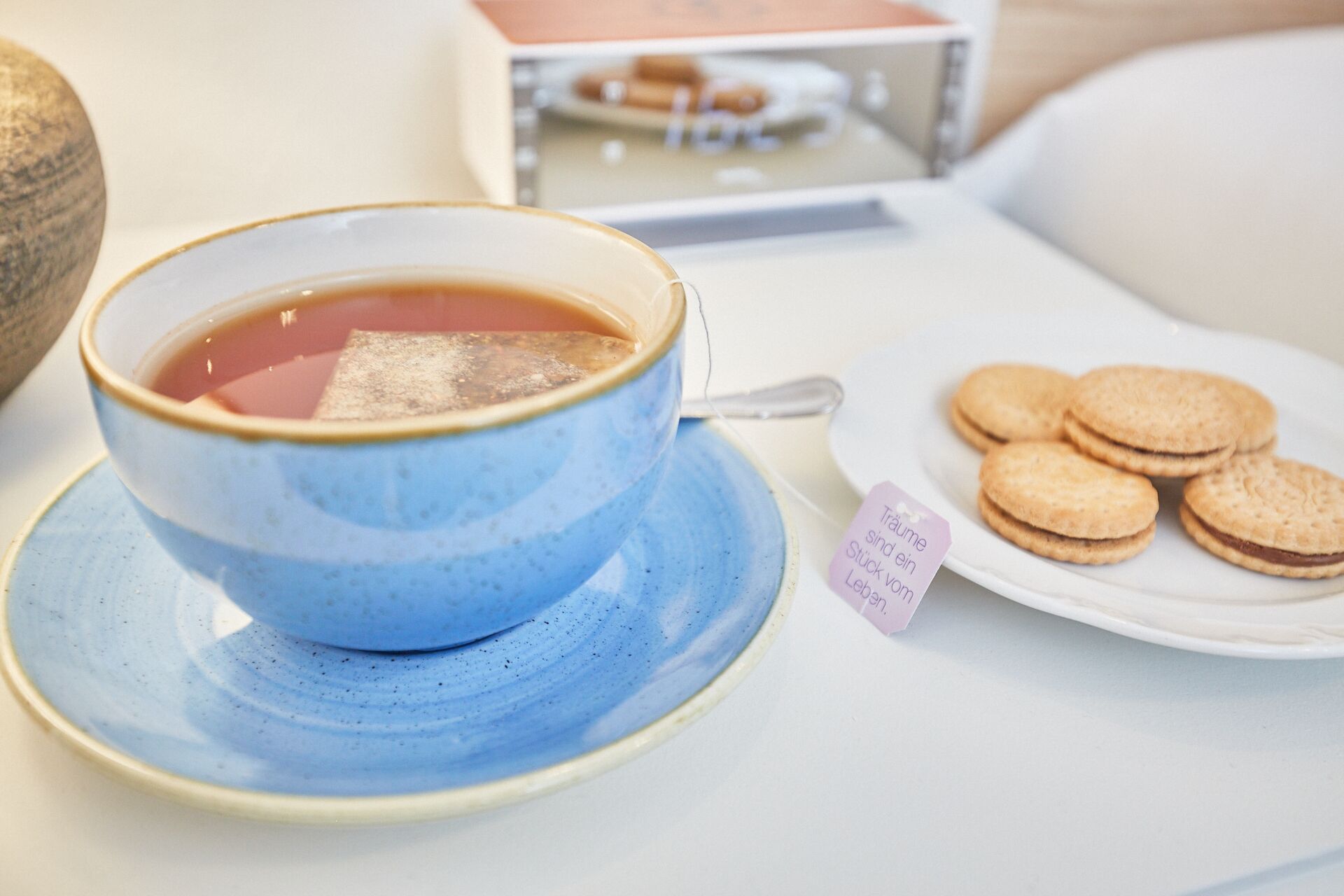 Eine blaue Tasse Tee mit Untertasse, ein Teller Kekse und eine kleine Schachtel im Hintergrund.