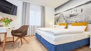 A hotel room with a bed, a desk with a chair, a TV, and a painting on the wall.