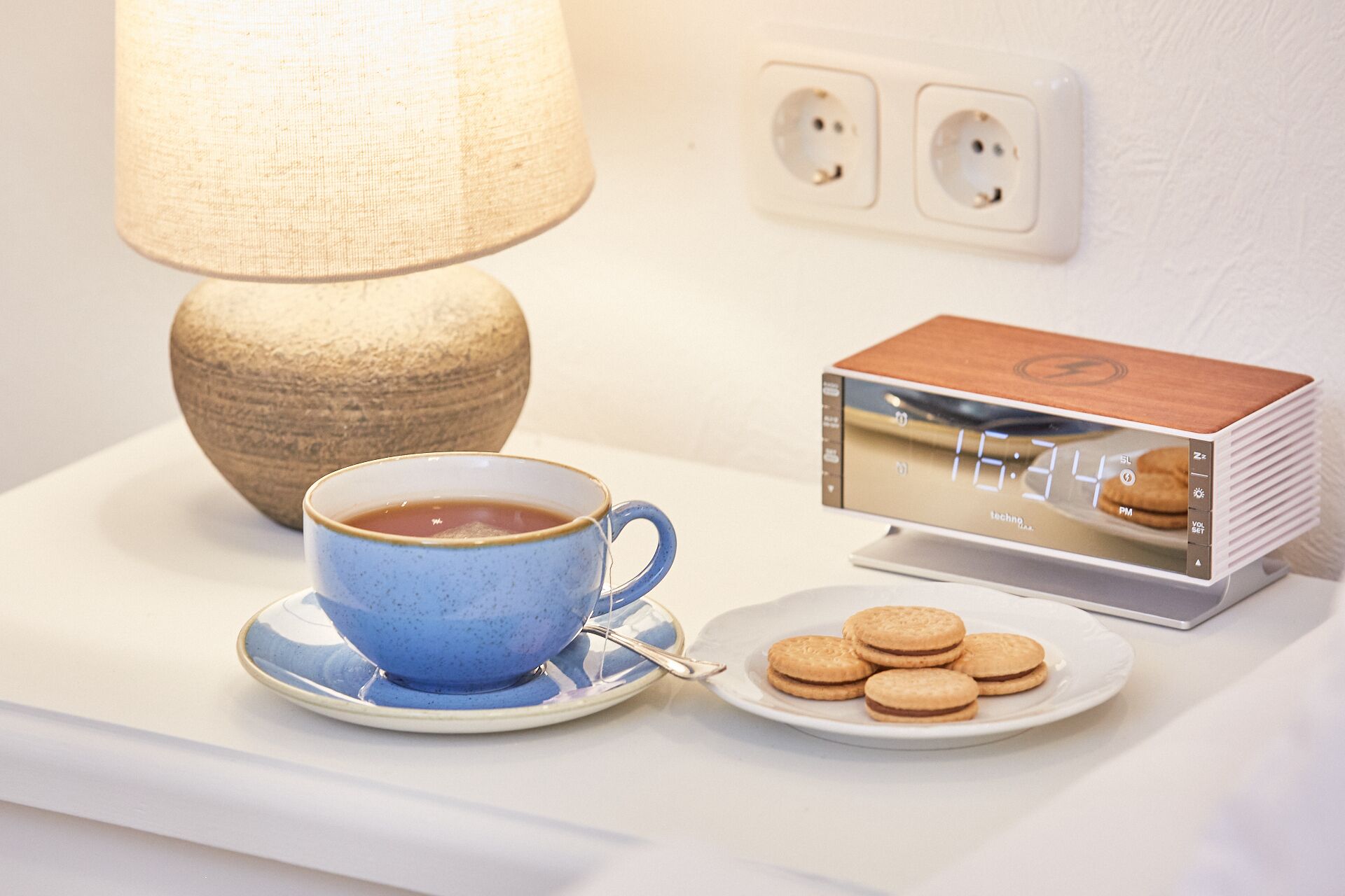 A table with a blue cup of coffee, a plate of cookies, a lamp, and a digital clock.