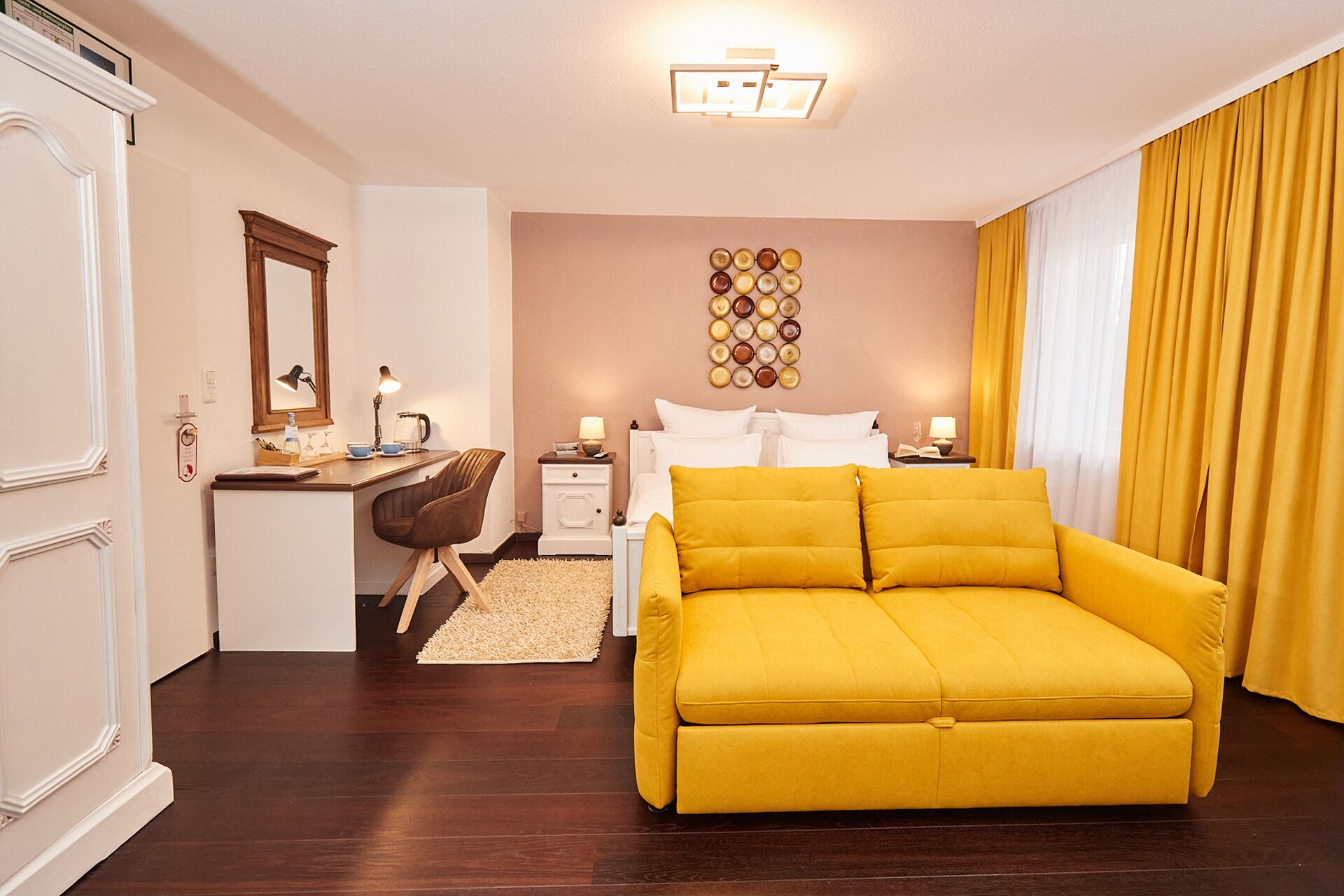A hotel room with a yellow sofa, a desk with a lamp, a bed with white linens, and a wall with a circular pattern.
