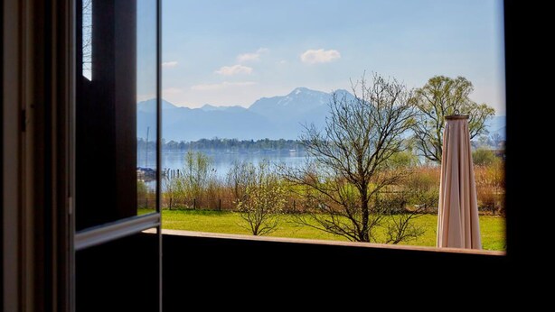 A view through a window of a lake with mountains in the background, a tree, and a vase on the windowsill. - Zum Fischer am See (Prien am Chiemsee)