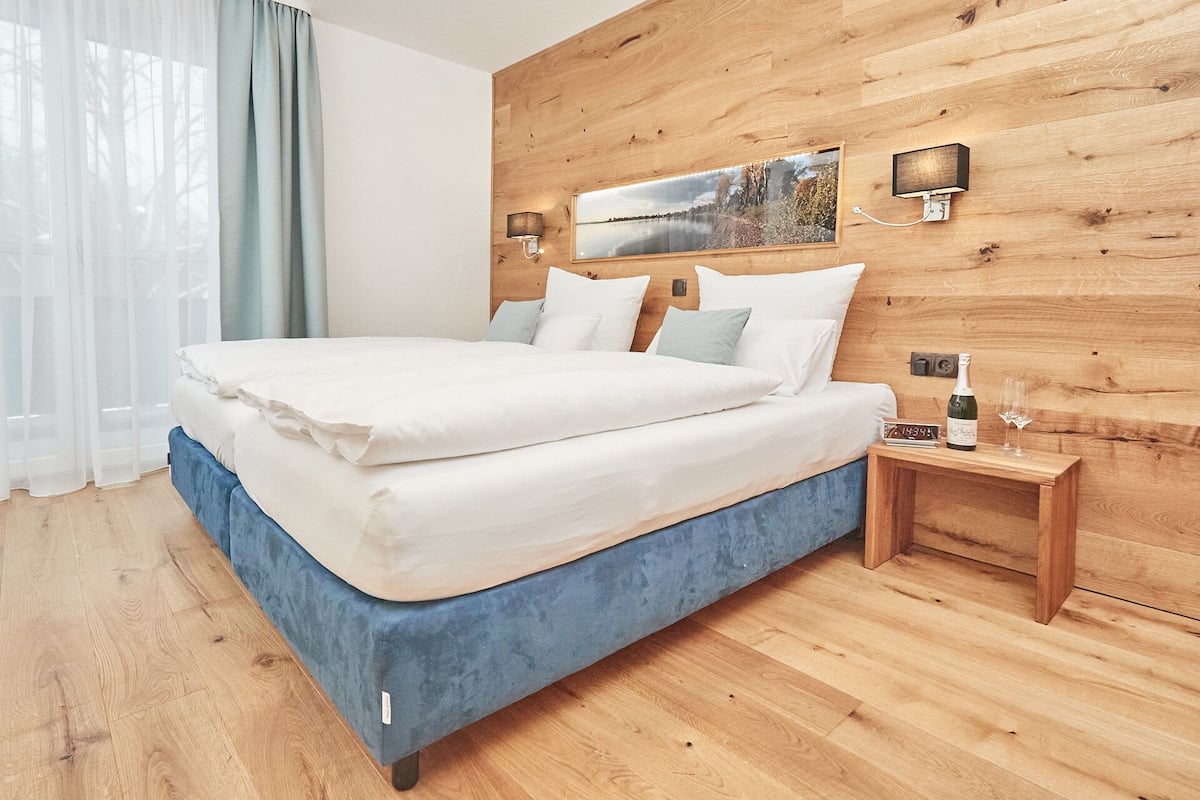 A bedroom with a wooden headboard, a bed with white bedding, a nightstand with a bottle and glasses, and a clock.