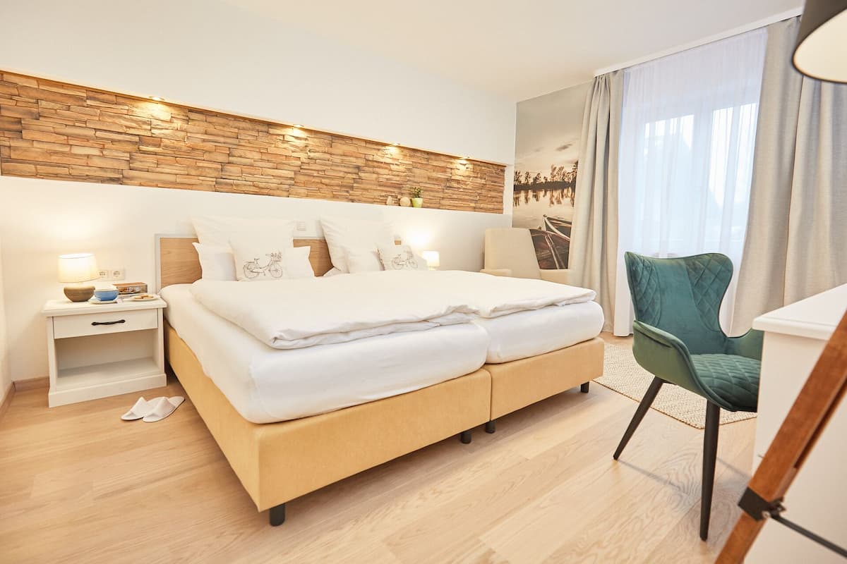 A modern hotel room with a wooden headboard, a large bed with white linens, a green armchair, and a view of a bridge through the window.