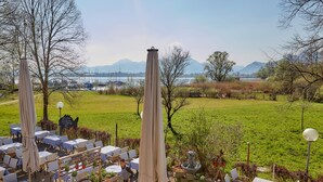 An outdoor dining area with tables and chairs, a fountain, and a view of the lake and mountains in the background. - Zum Fischer am See (Prien am Chiemsee)