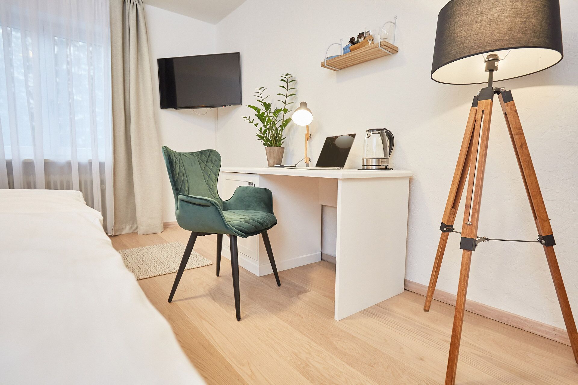 A hotel room with a bed, a desk with a laptop, a chair, a floor lamp, and a TV on the wall.