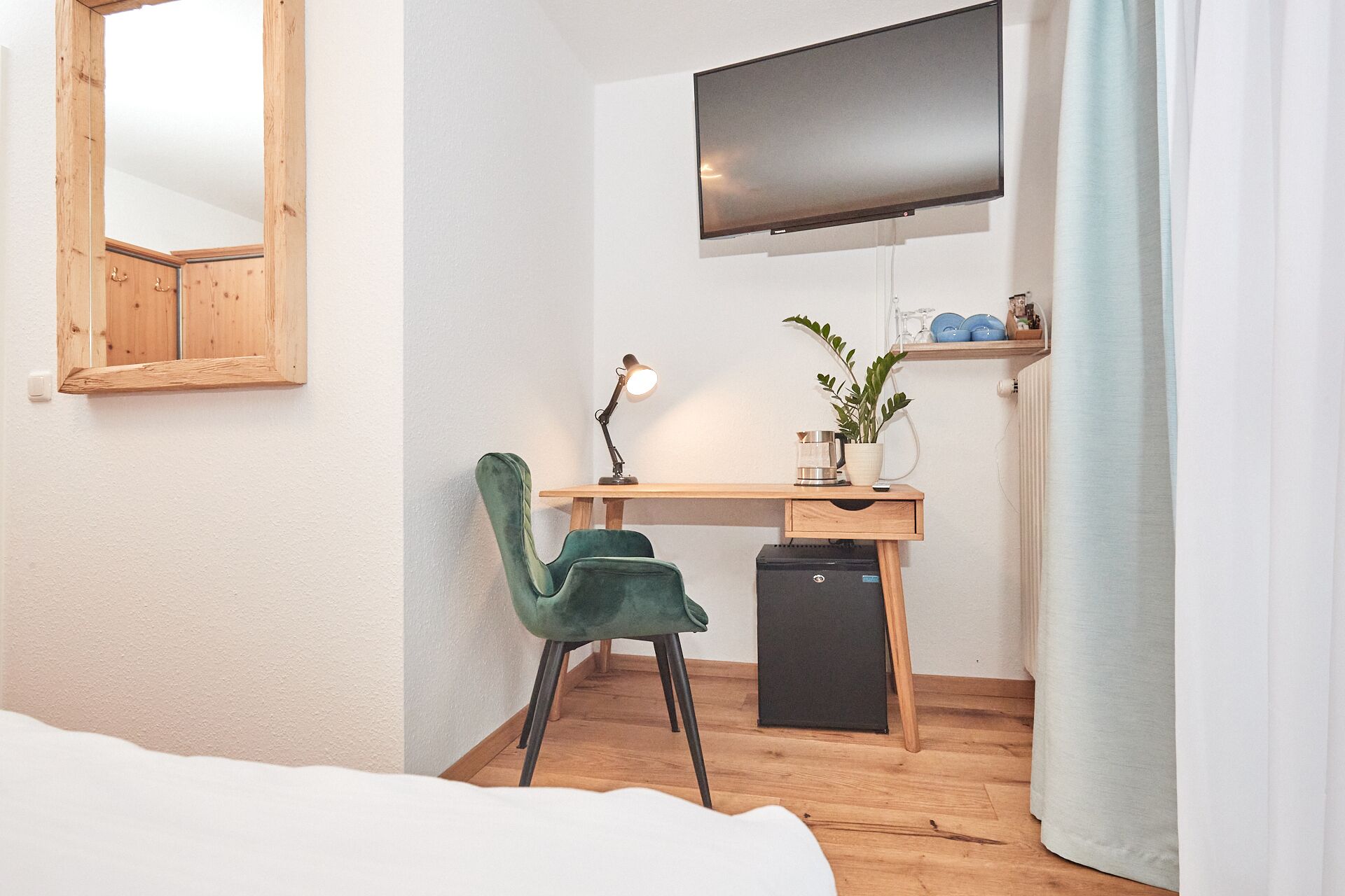 Ein Schlafzimmer mit Bett, Schreibtisch mit Stuhl, Fernseher, Lampe, Zimmerpflanze und einer Regalreihe mit Geschirr.