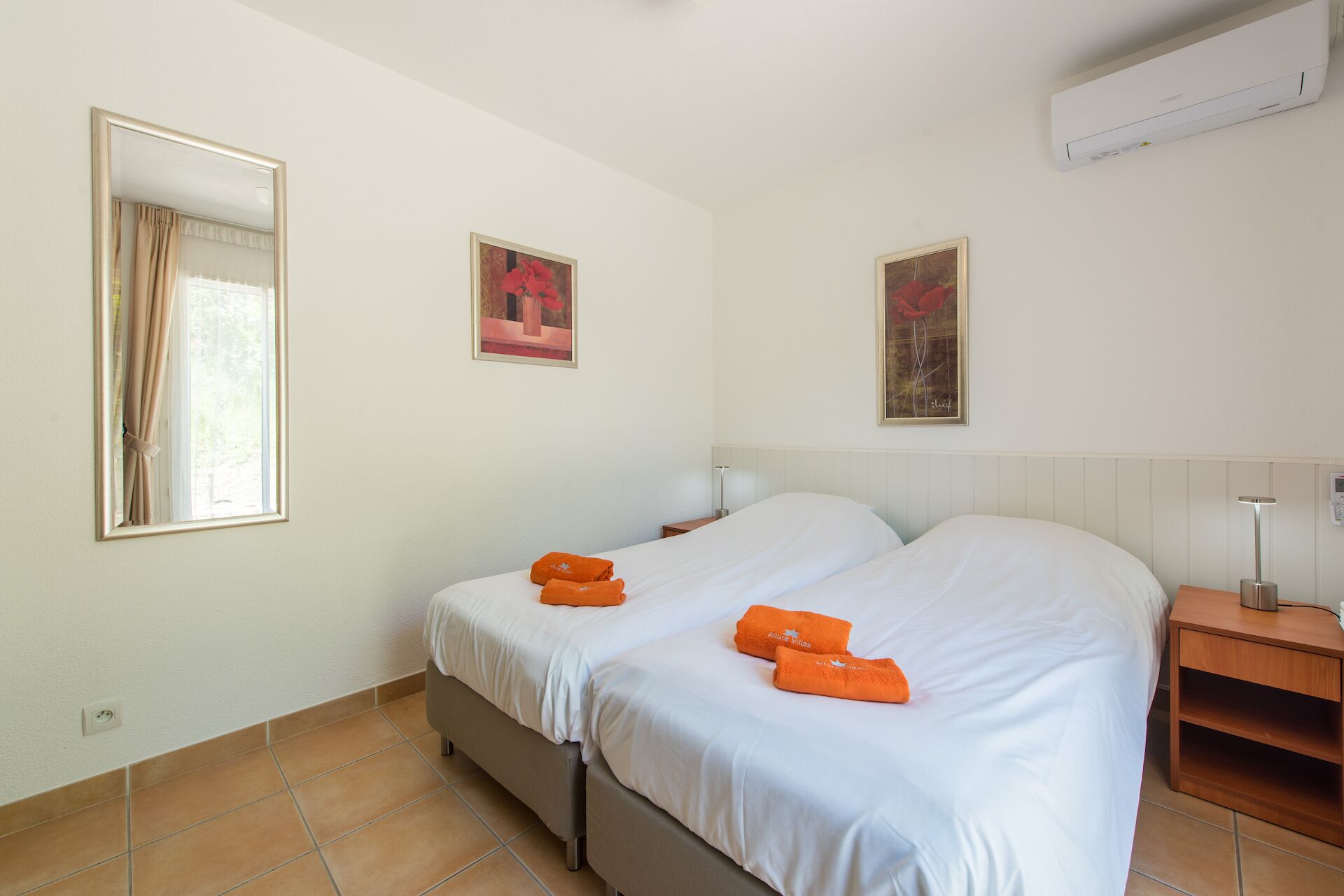 Habitación de hotel con dos camas, una mesita de noche, una lámpara y dos cuadros enmarcados en la pared.