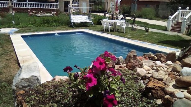 A rectangular swimming pool with a stone border, surrounded by a grassy area with a white fence and a few chairs.