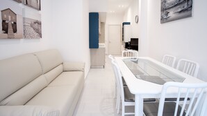 Una sala de estar moderna con un sofá beige, una mesa de comedor con cubierta de vidrio y sillas blancas, y obras de arte en las paredes.