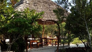 Ein Gazebo mit einem Reetdach, Holzbanken und einem gepflasterten Weg, umgeben von üppiger Vegetation.