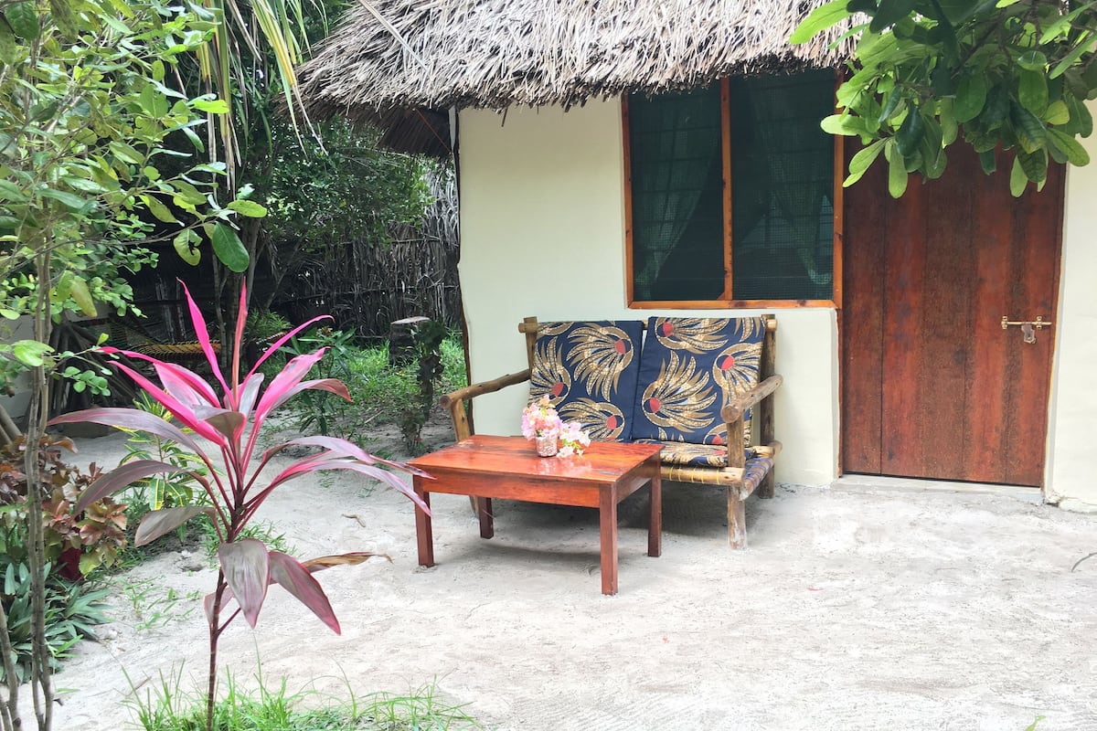 Uma cabana com telhado de palha, uma porta de madeira, uma poltrona acolchoada com padrão floral e uma pequena mesa com um vaso de flores.