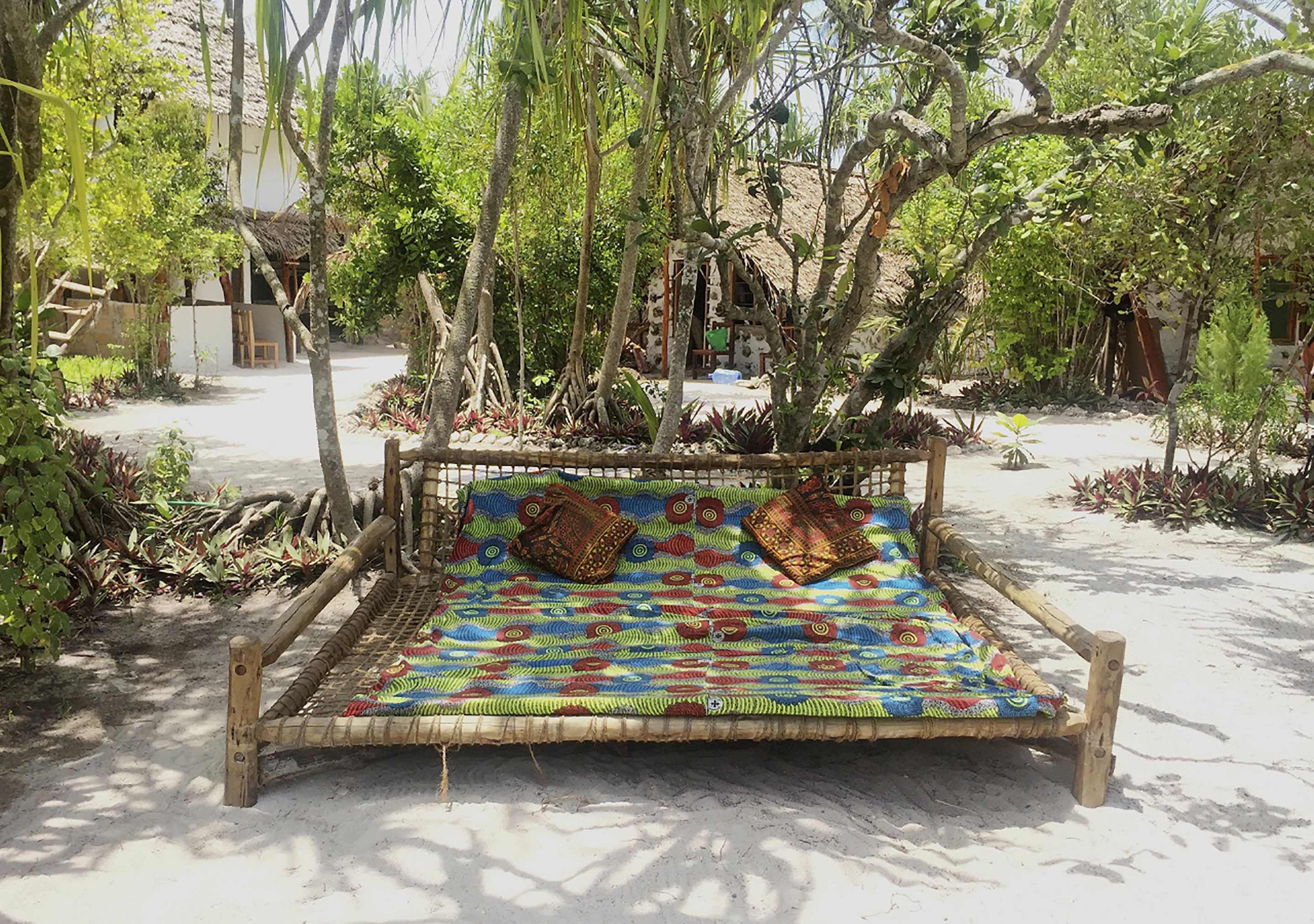 Una cama de madera rústica con un colorido cobijo tejido y dos almohadas, ubicada en un jardín natural y lleno de árboles.