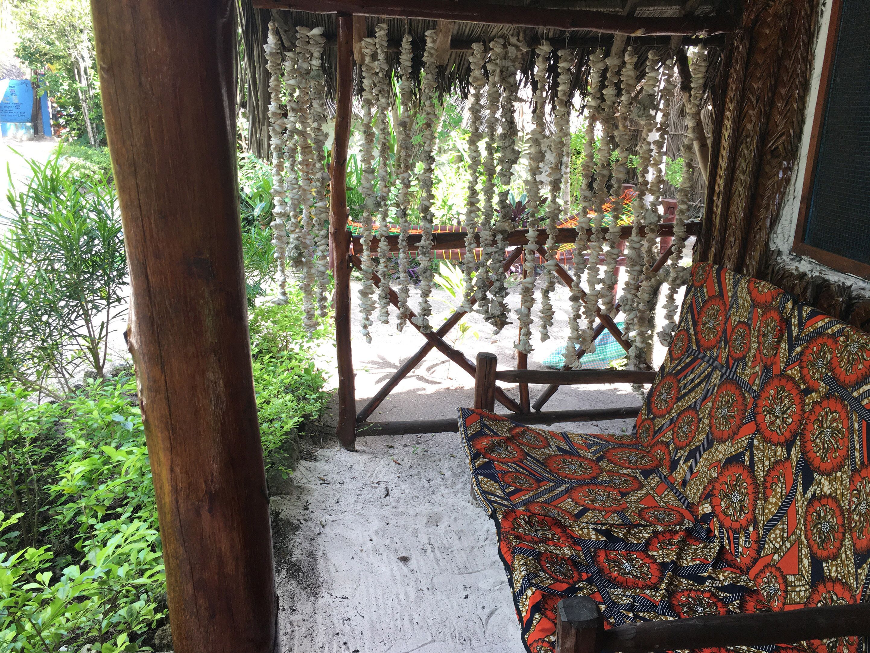 Un banc en bois avec un coussin à motifs, une structure surmontée d’un toit de chaume et des coquillages suspendus.