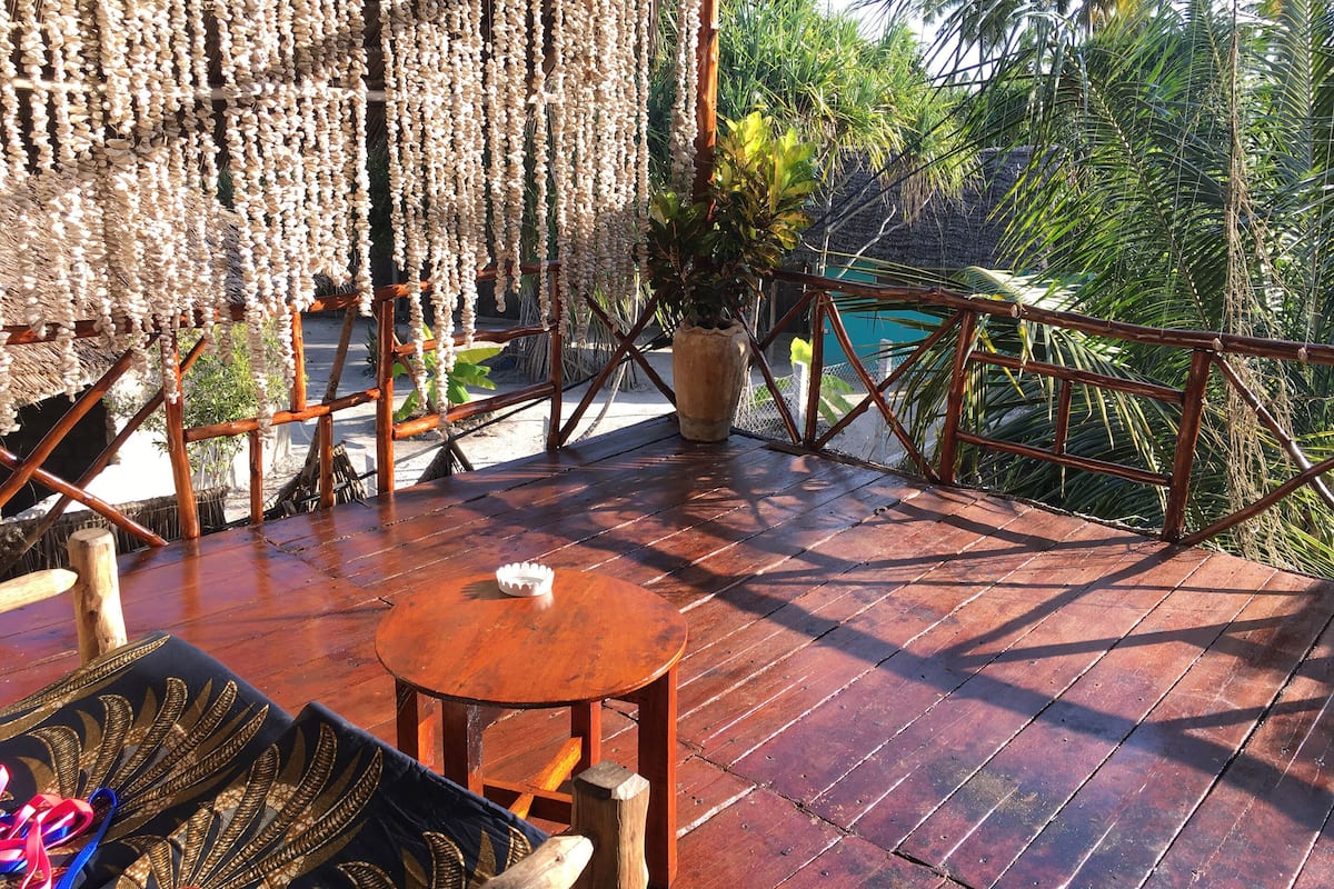 Um deck de madeira com mesa redonda e cadeiras, cercado por um cercado de bambu e plantas em vasos.