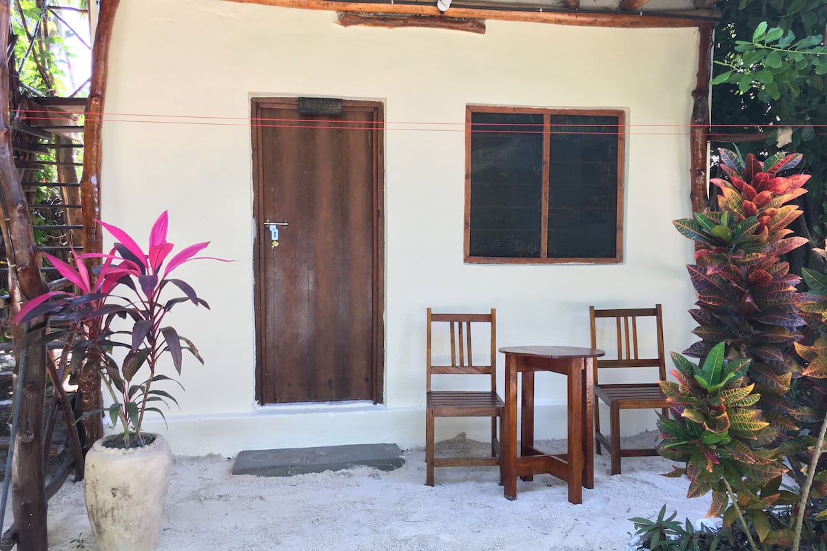 Um pequeno edifício térreo com porta e janela de madeira, rodeado por plantas e um espaço de estar ao ar livre simples.