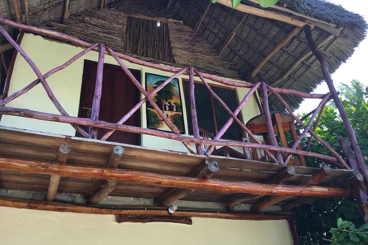Uma estrutura de madeira tradicional com telhado de palha, varanda com corrimão de madeira e porta que dá para um espaço interno.
