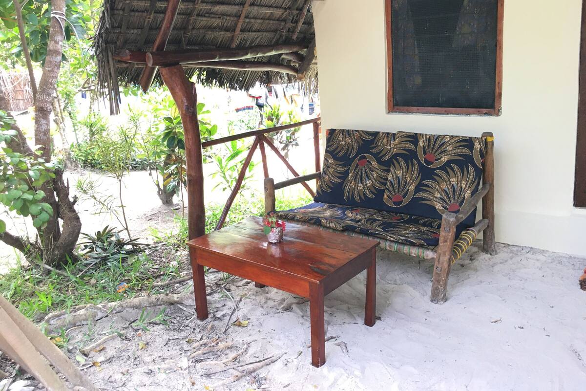 Um banco de madeira com almofada estampada, uma mesa pequena e uma estrutura de telhado de palha.
