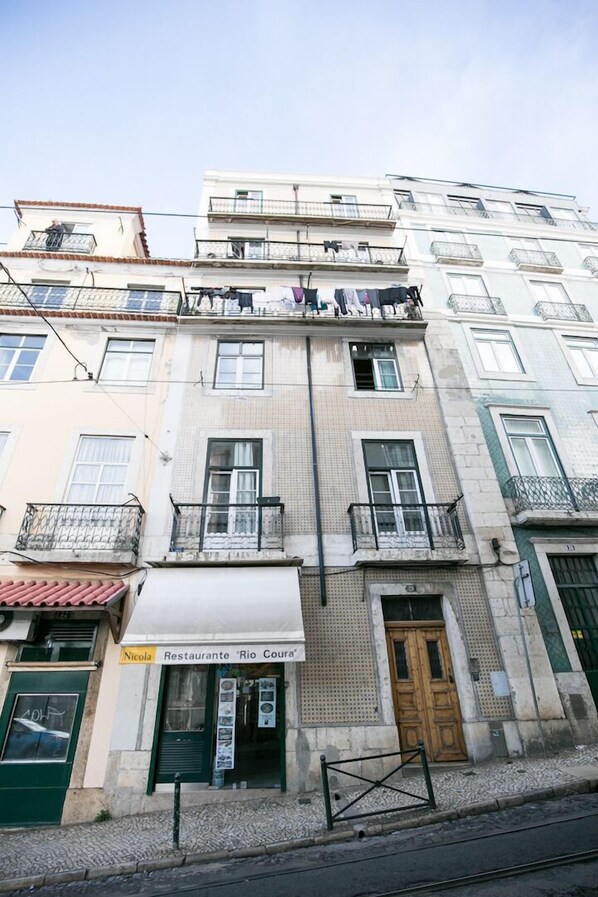 Um edifício com vários andares e varandas, um restaurante chamado "Nicola Restaurante Rio Coura" no térreo, e uma porta verde.