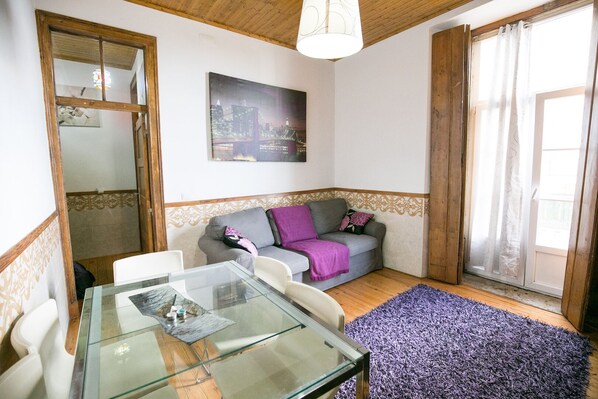A cozy living area with a glass-top table, a grey sofa with a purple throw, and a cityscape painting on the wall.