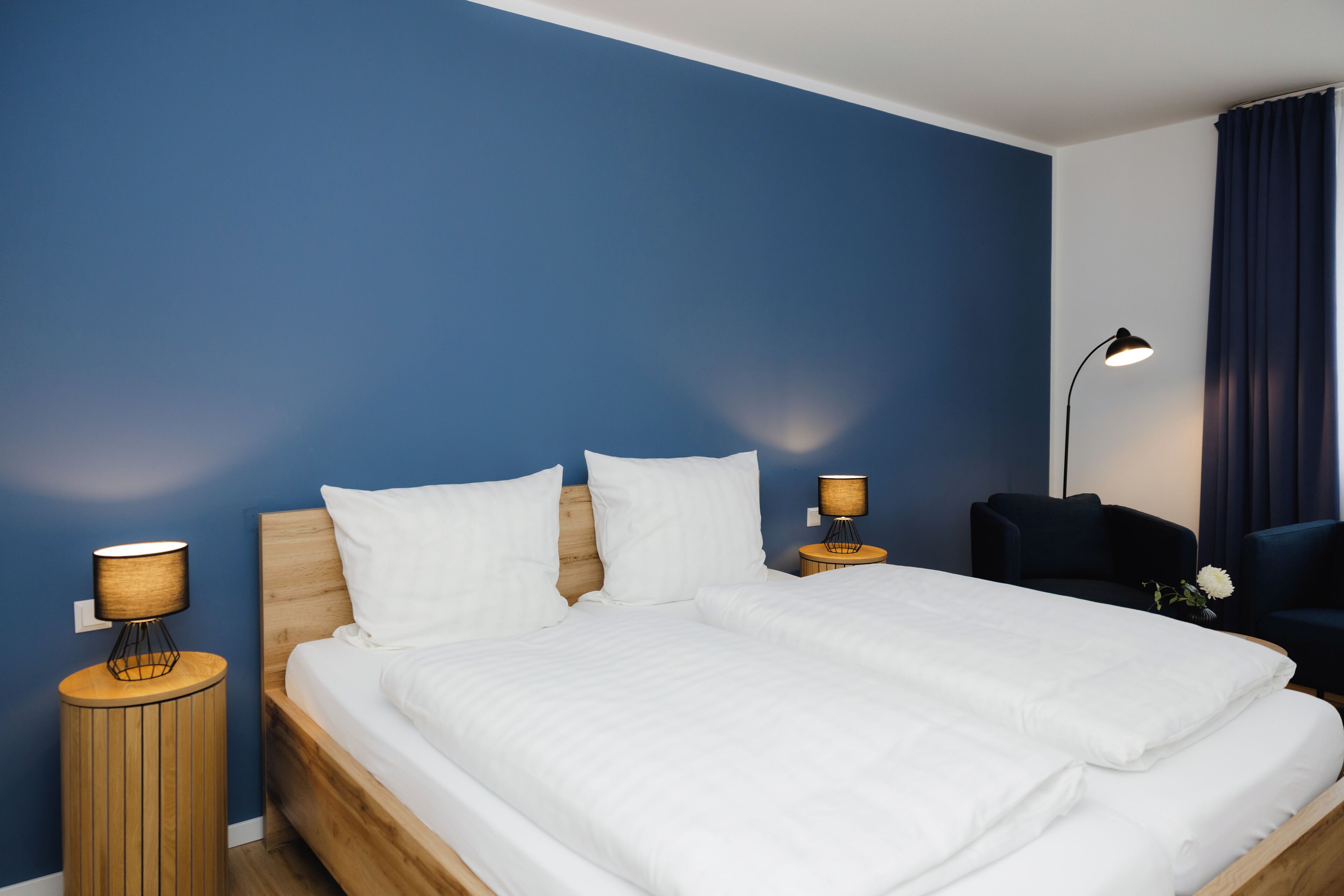 Una cama doble con ropa blanca, dos mesitas de noche con lámparas, una pared azul oscuro y una silla azul oscuro.