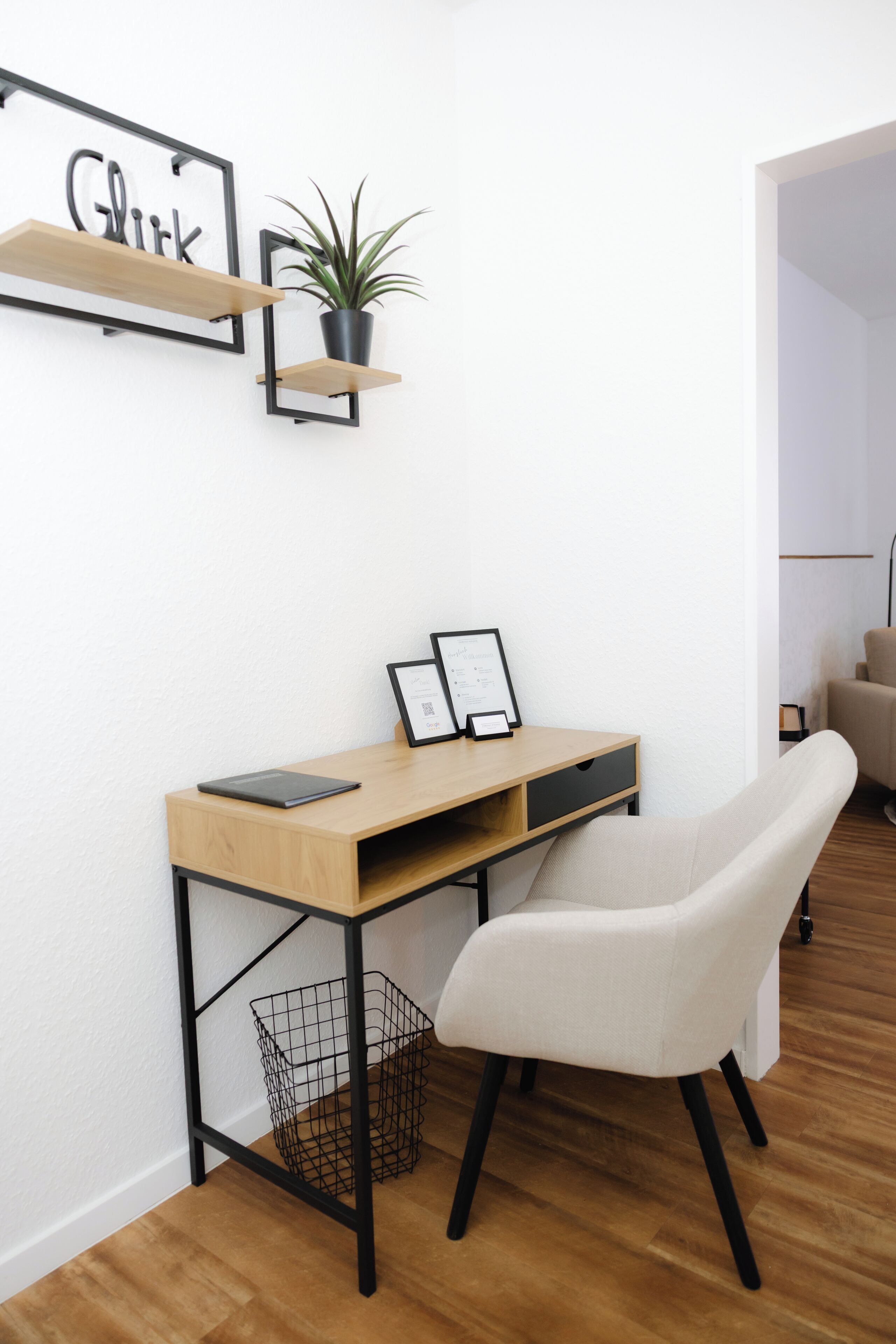 Un espacio de oficina moderno con un escritorio de madera, una silla blanca, una cesta de metal negro y una planta en maceta sobre una estantería negra.