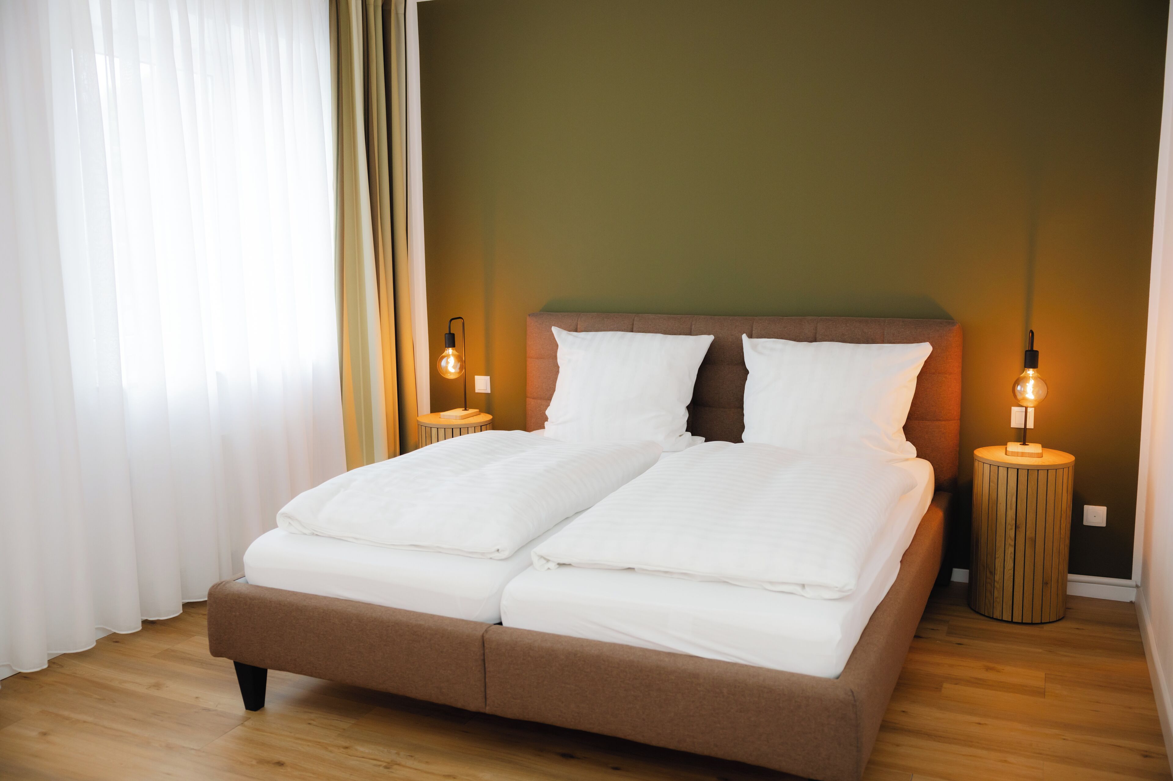 A double bed with white linens, two bedside tables with lamps, a wooden floor, and a green wall.