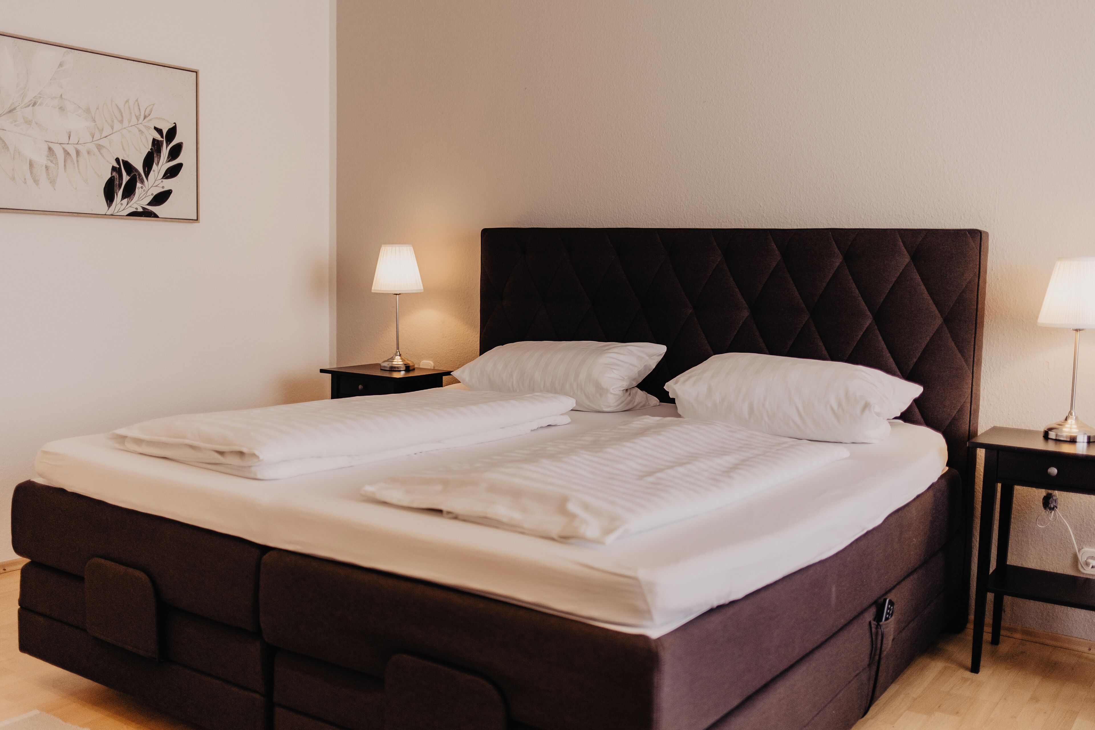 Ein Doppelbett mit dunklem Kopfteil, zwei Nachttische mit Lampen und ein gerahmtes Kunstwerk an der Wand.