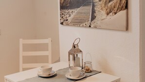 Un comedor con una mesa blanca, dos sillas, un cuadro de una escena de playa y un pequeño farol sobre la mesa.