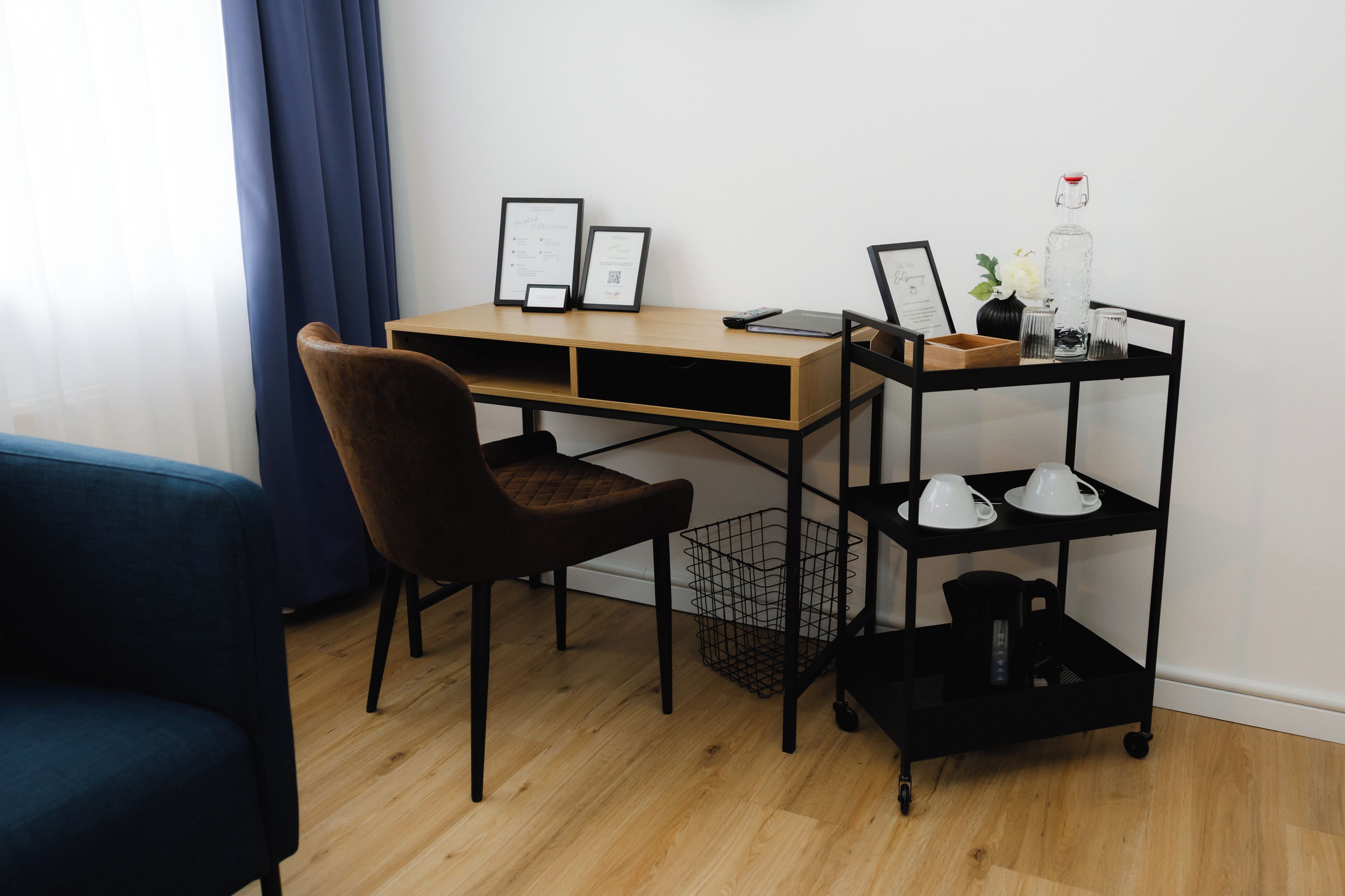 A modern workspace with a wooden desk, a brown chair, a black metal shelf with cups and a kettle, and framed certificates on the wall.