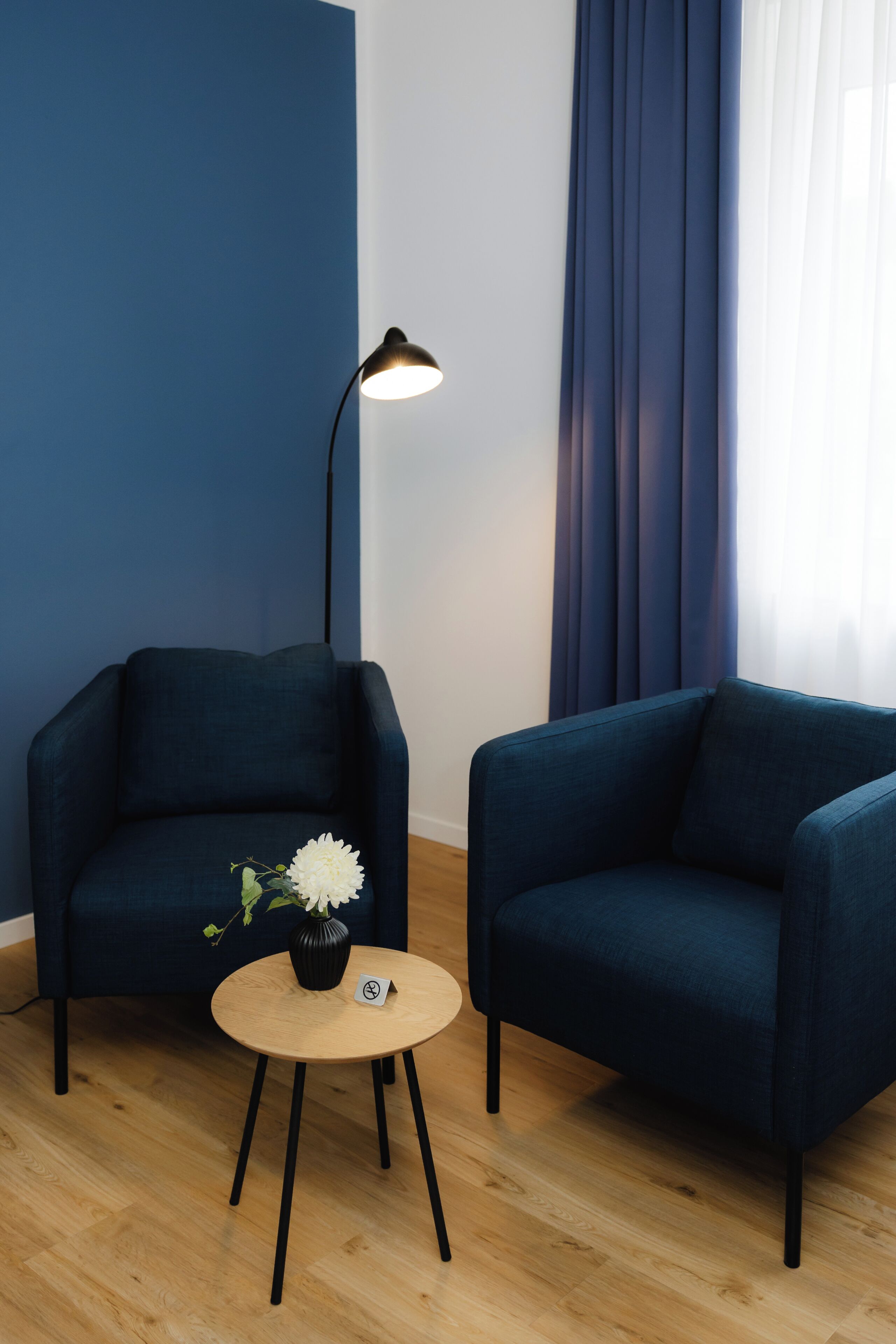Habitació moderna amb dues butaques blau fosc, una taula rodona de fusta amb un ram de flors, una làmpada de peu i un gran finestral amb cortines.