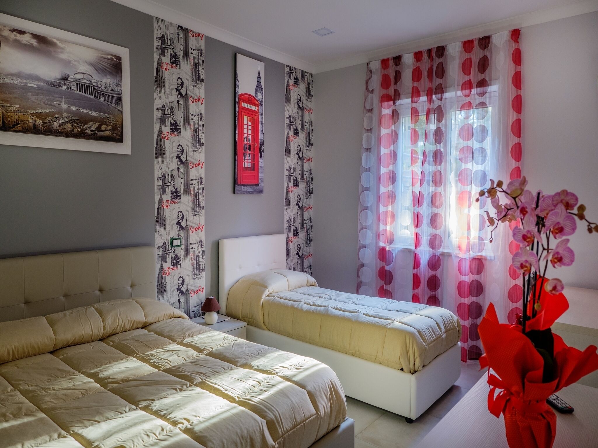 A bedroom with two beds, a large window with pink curtains, and a wall with cityscape wallpaper.