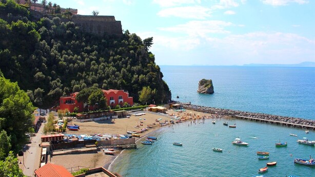 A coastal town with a beach, boats, and a historic fort on a hill.