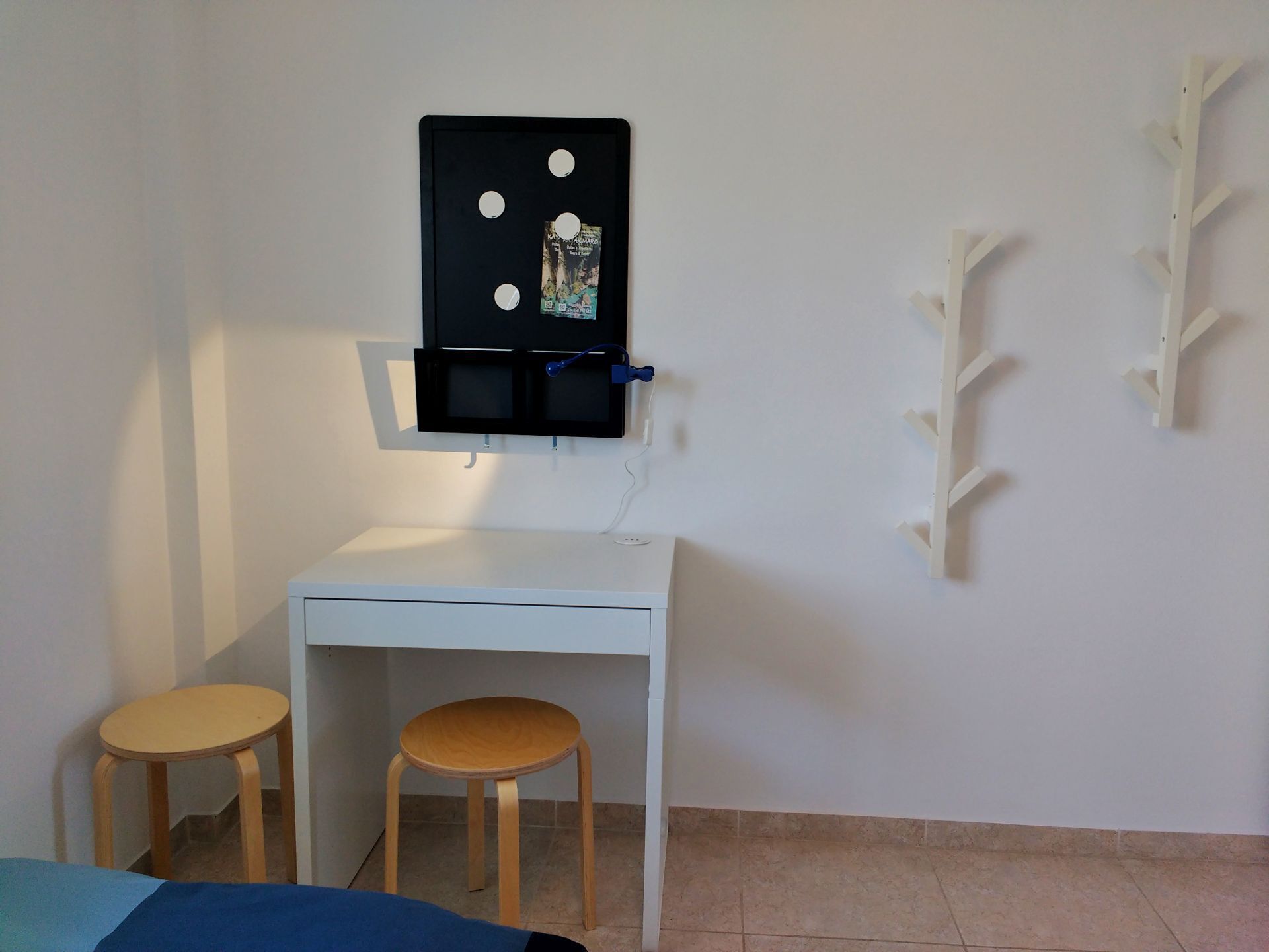 Um quarto pequeno com uma escrivaninha branca, dois bancos de madeira e uma TV fixada na parede.