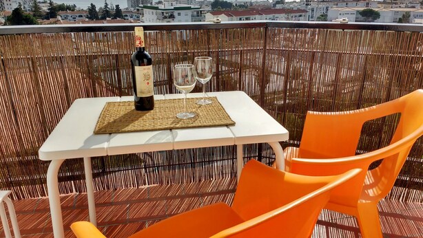 A balcony with a table, two orange chairs, a bottle of wine, and two glasses.