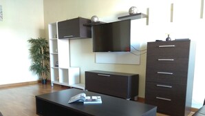 Een moderne woonkamer met een flatscreen televisie aan de muur, een donker houten kast met lades, een zwarte salontafel met tijdschriften, en een potplant.