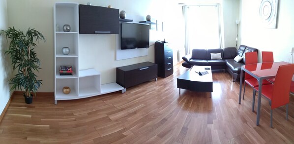 A modern living room with a flat-screen TV, a sofa, a coffee table, and a dining area with red chairs.