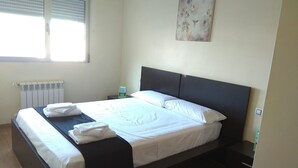 Een net opgemaakt bed met witte lakens en een donker hoofdbord, in een kamer met een radiator, een klein nachtkastje en een wanddecoratie.