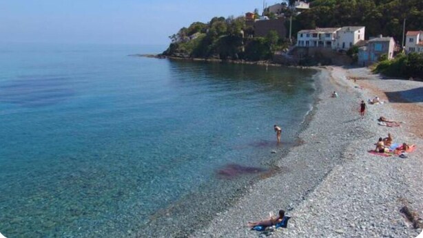A pebble beach with clear water, people sunbathing, and buildings on a hillside.