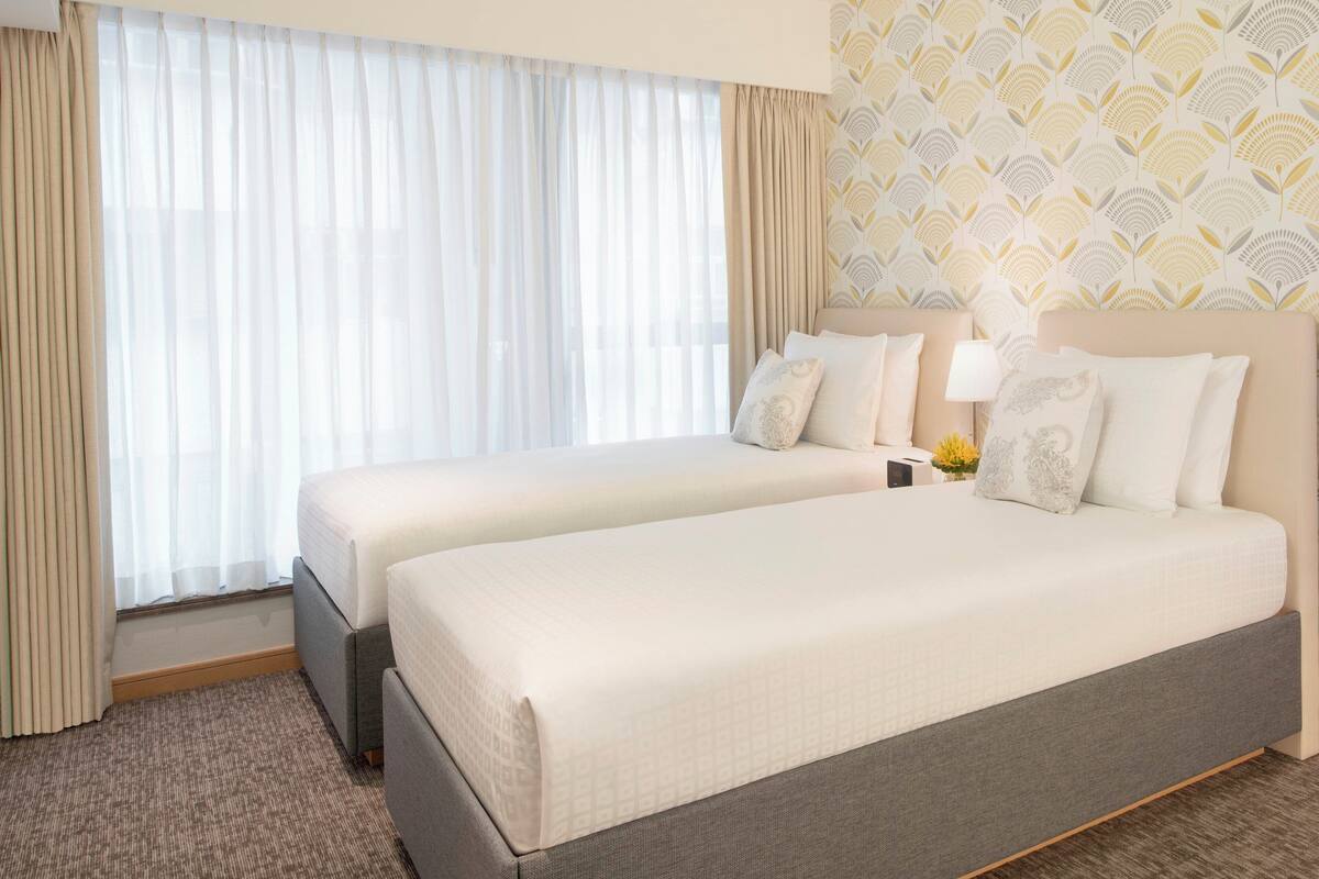 A hotel room with two beds, a large window with sheer curtains, a patterned wallpaper, and a small table with a lamp.