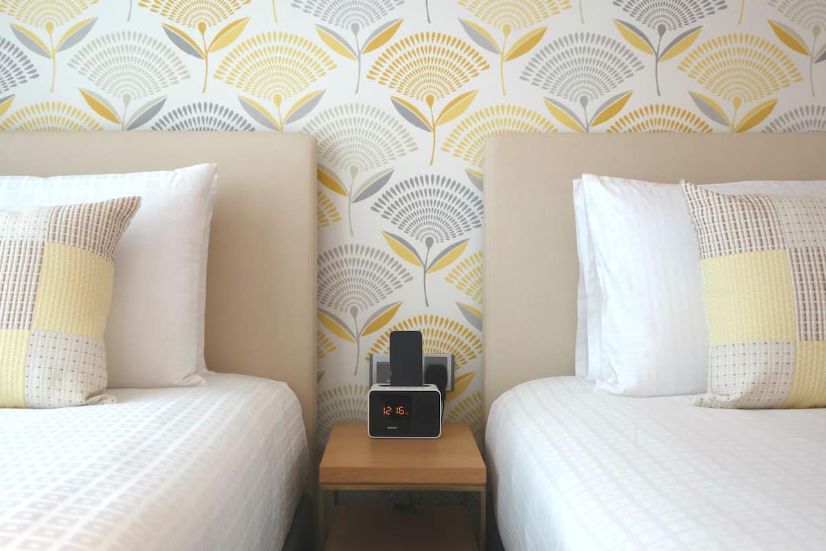 A hotel room with two beds, a nightstand with a digital clock, and a patterned wallpaper.