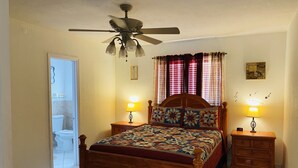 Une chambre à coucher avec un lit en bois, deux tables de chevet avec des lampes, un ventilateur de plafond et une vue sur la salle de bain.