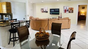 A modern living room with a glass dining table, leather sofa, and a TV mounted on the wall.