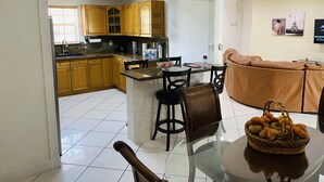 A modern kitchen with wooden cabinets, a dining area with a glass table, and a living area with a brown sofa.