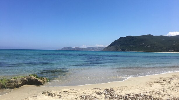 Une plage avec une eau d’un bleu limpide, un rivage sablonneux et une montagne en arrière-plan.